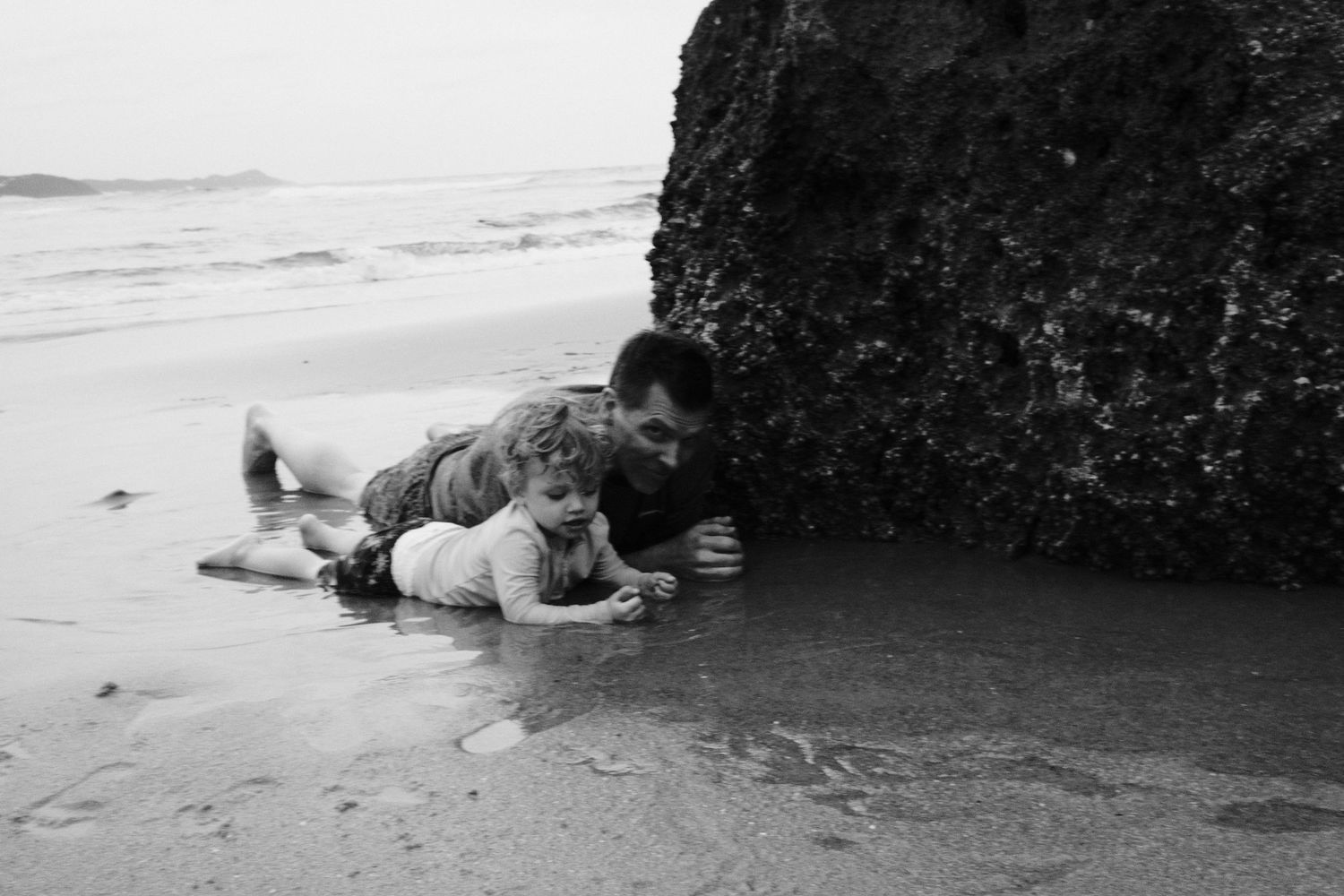 Man and child lying on wet sand near a large rock on a beach in black and white photography.