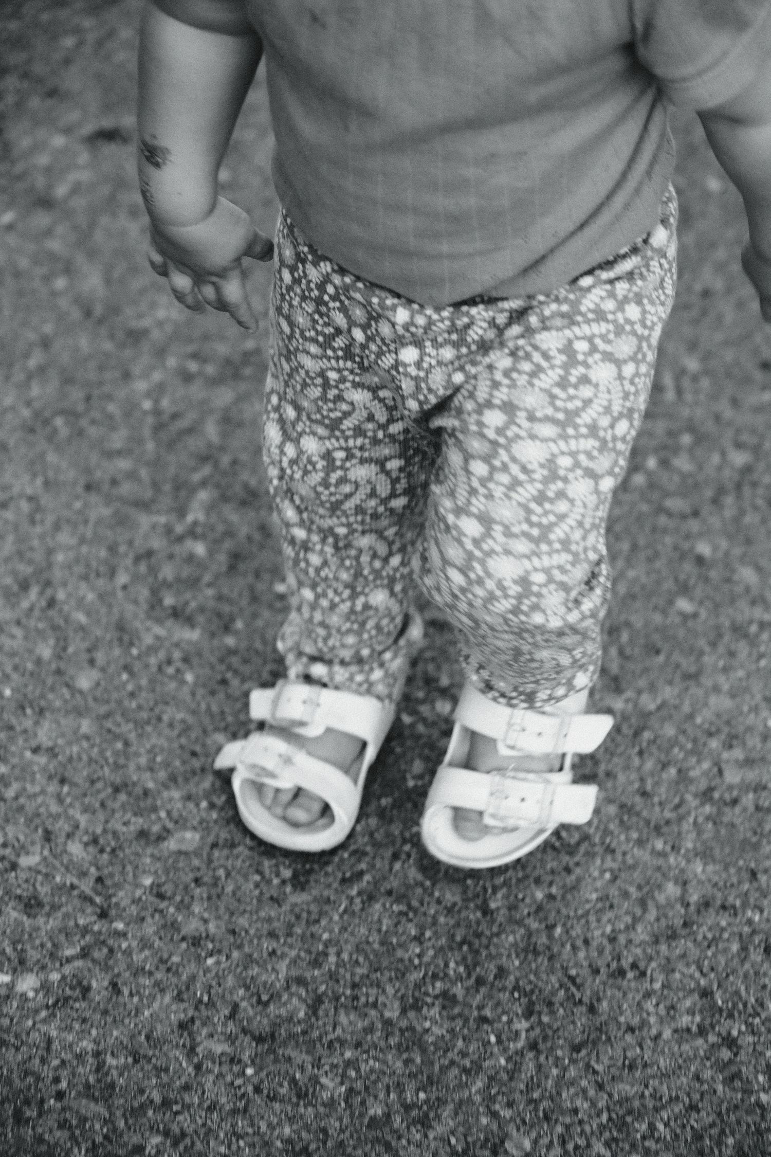Black and white close-up of a baby's feet wearing white sandals and patterned leggings.