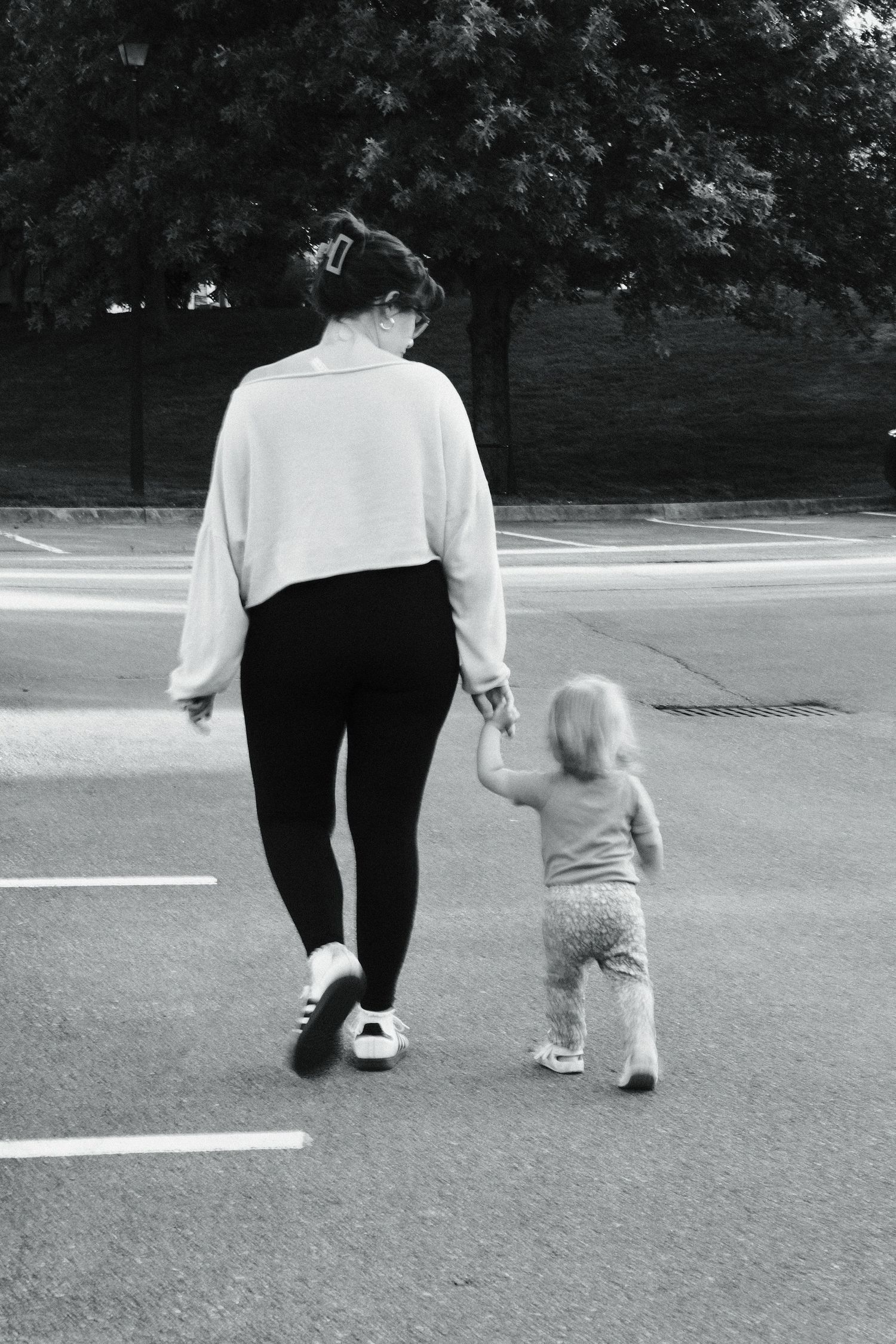 Black and white photo of a mother and toddler walking hand in hand across a parking lot.