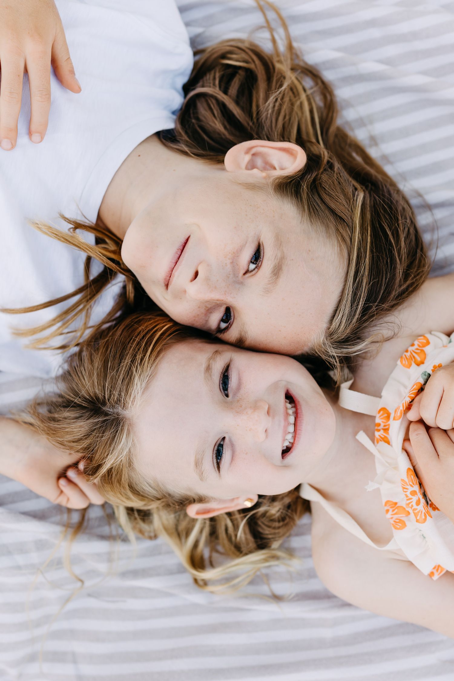 brother sister lying on white bedding with their heads close together while smiling at the camera.