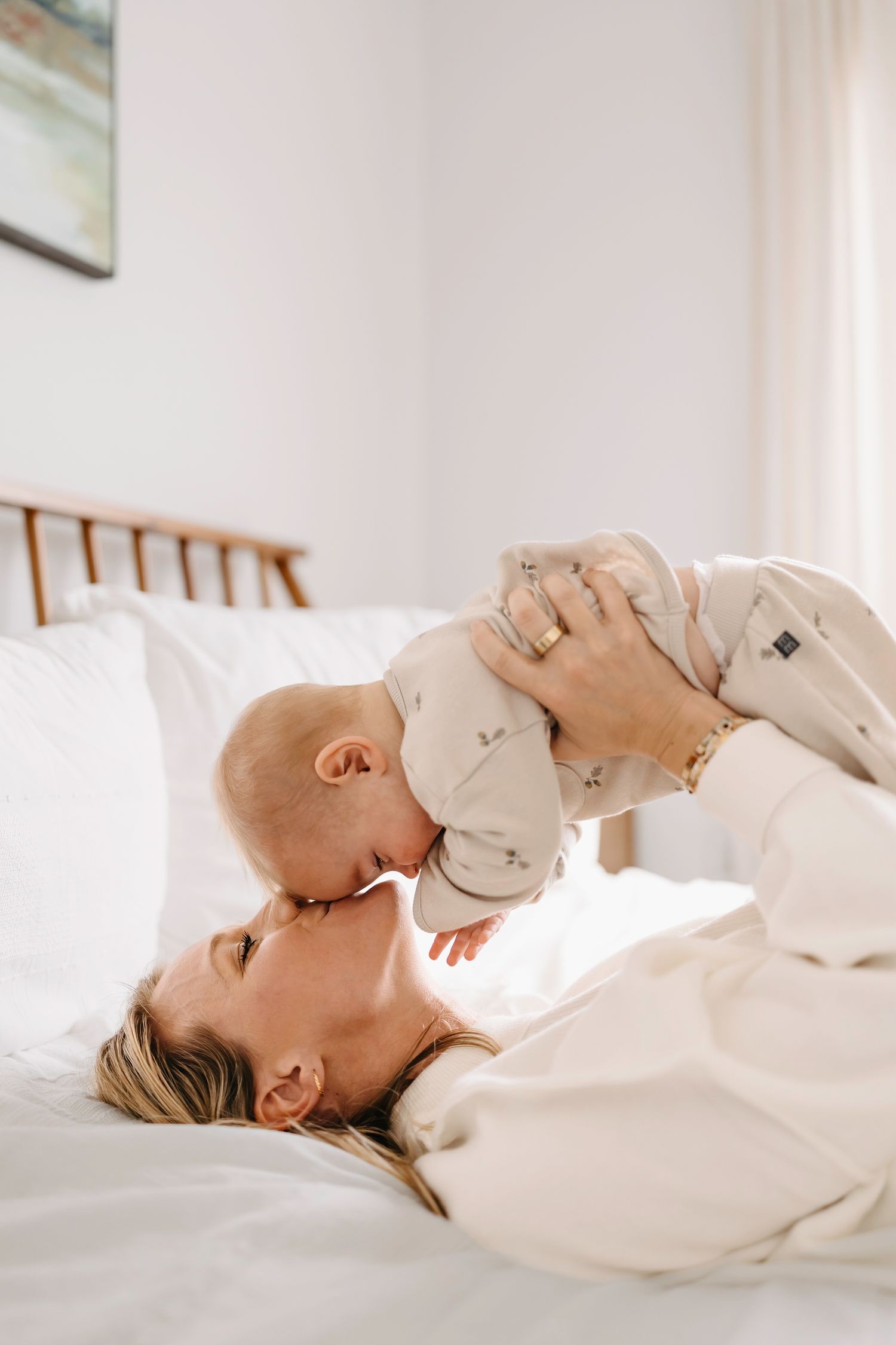 A parent in white clothing lies on bed holding baby up in air against bright window light.