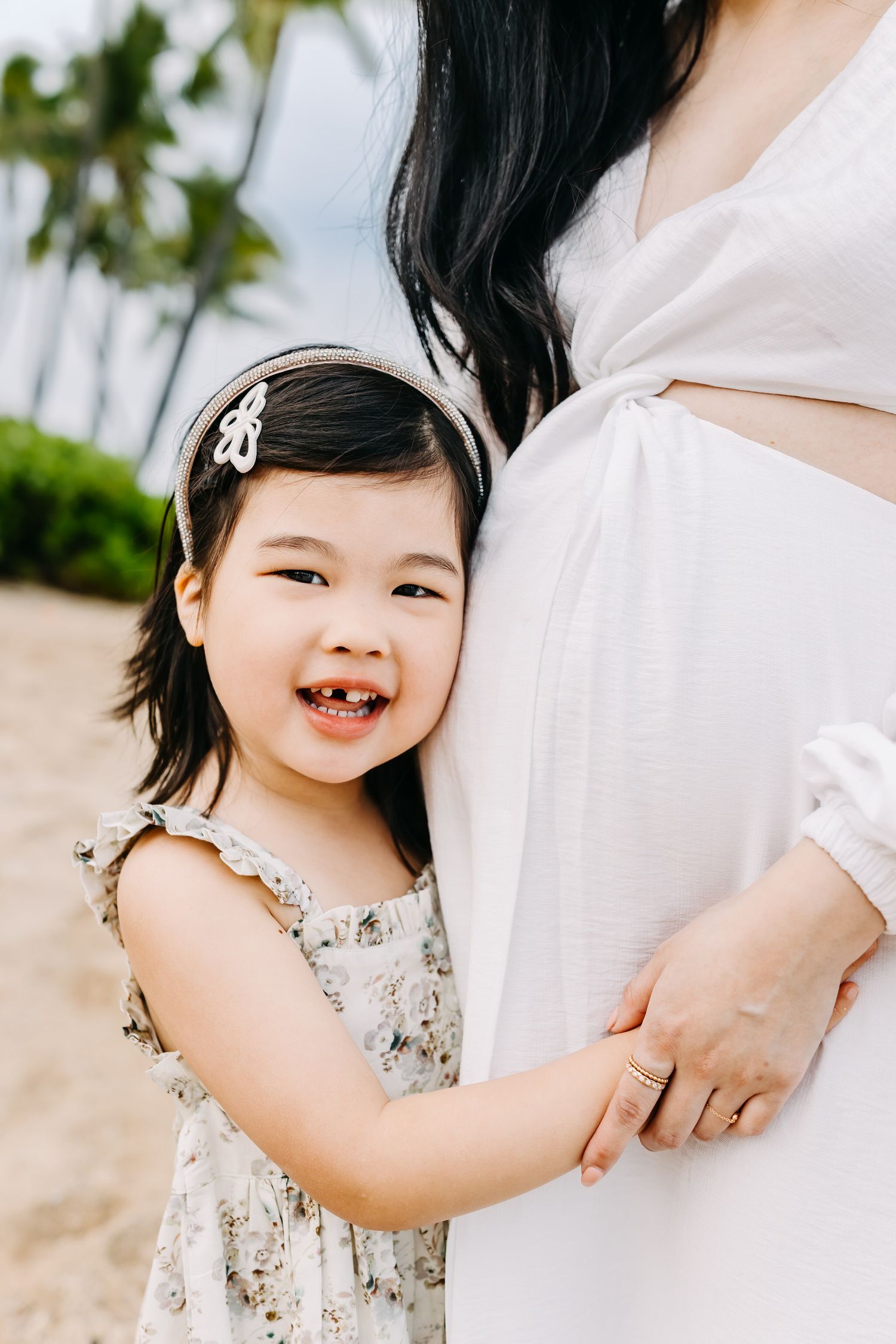 A small child in a floral dress smiles joyfully while holding an adult's hand at an outdoor location.