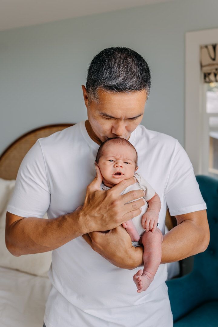 A dad in a white t-shirt holds and kisses a newborn baby in a series of tender moments on a couch.
