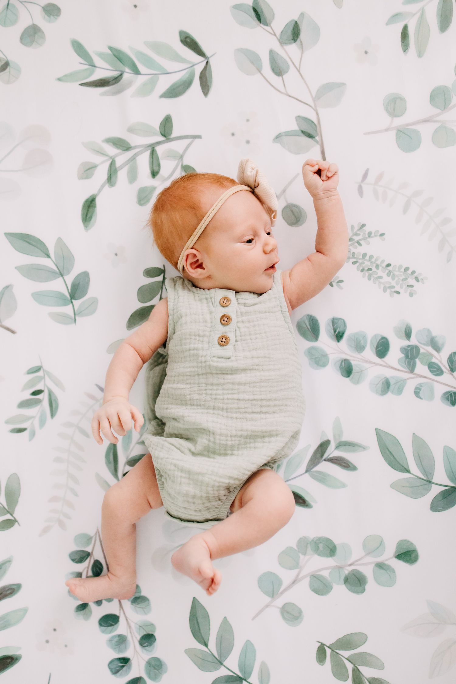 Baby wearing sage green romper lies on white blanket with delicate eucalyptus leaf pattern background.