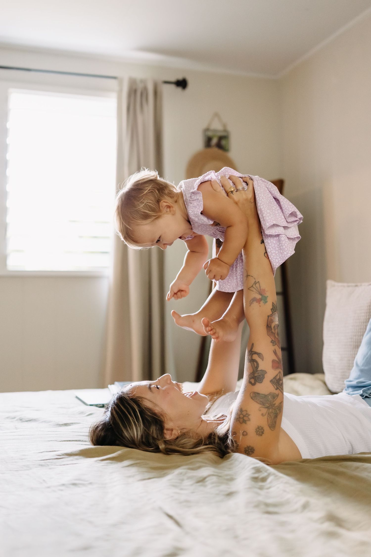 Parent lying on bed lifts baby wearing lavender outfit in playful bonding moment.