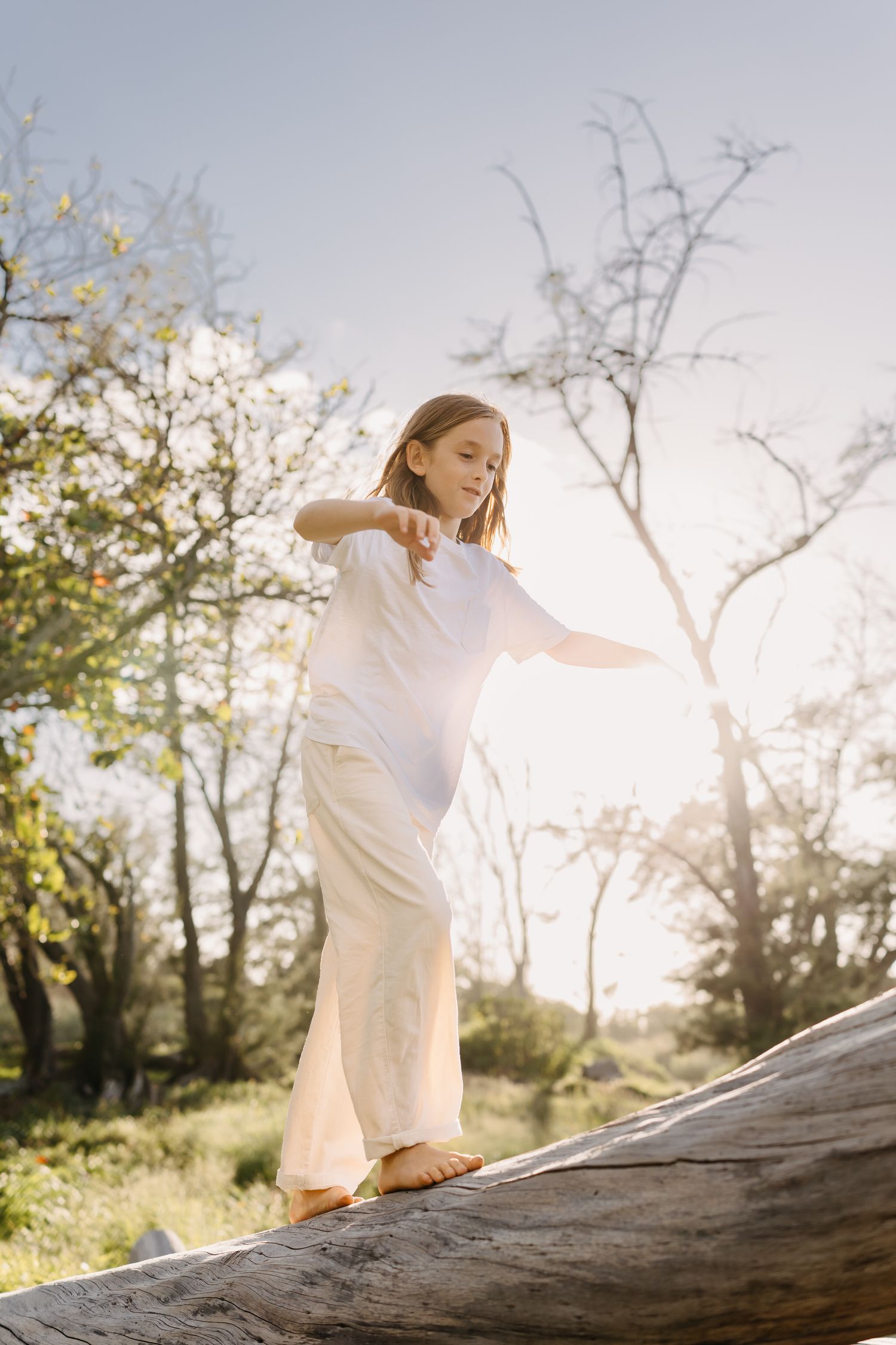 boy in a white dress gracefully balances on a fallen log in a sunlit forest during golden hour.