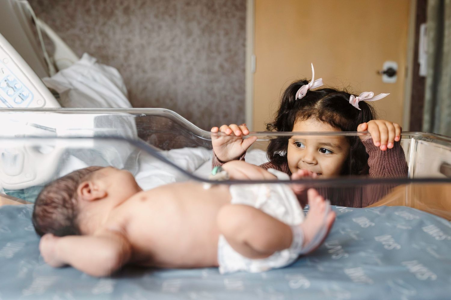 New sister looks through the clear bassinet at baby brother in hospital.