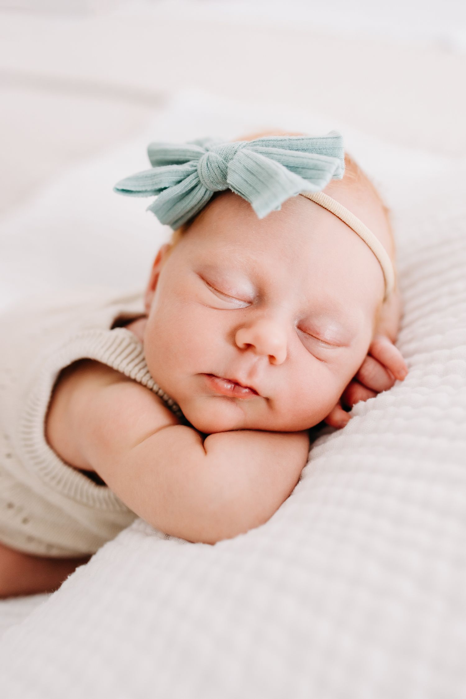 A sleeping newborn baby wearing a mint green bow headband rests peacefully on white bedding.