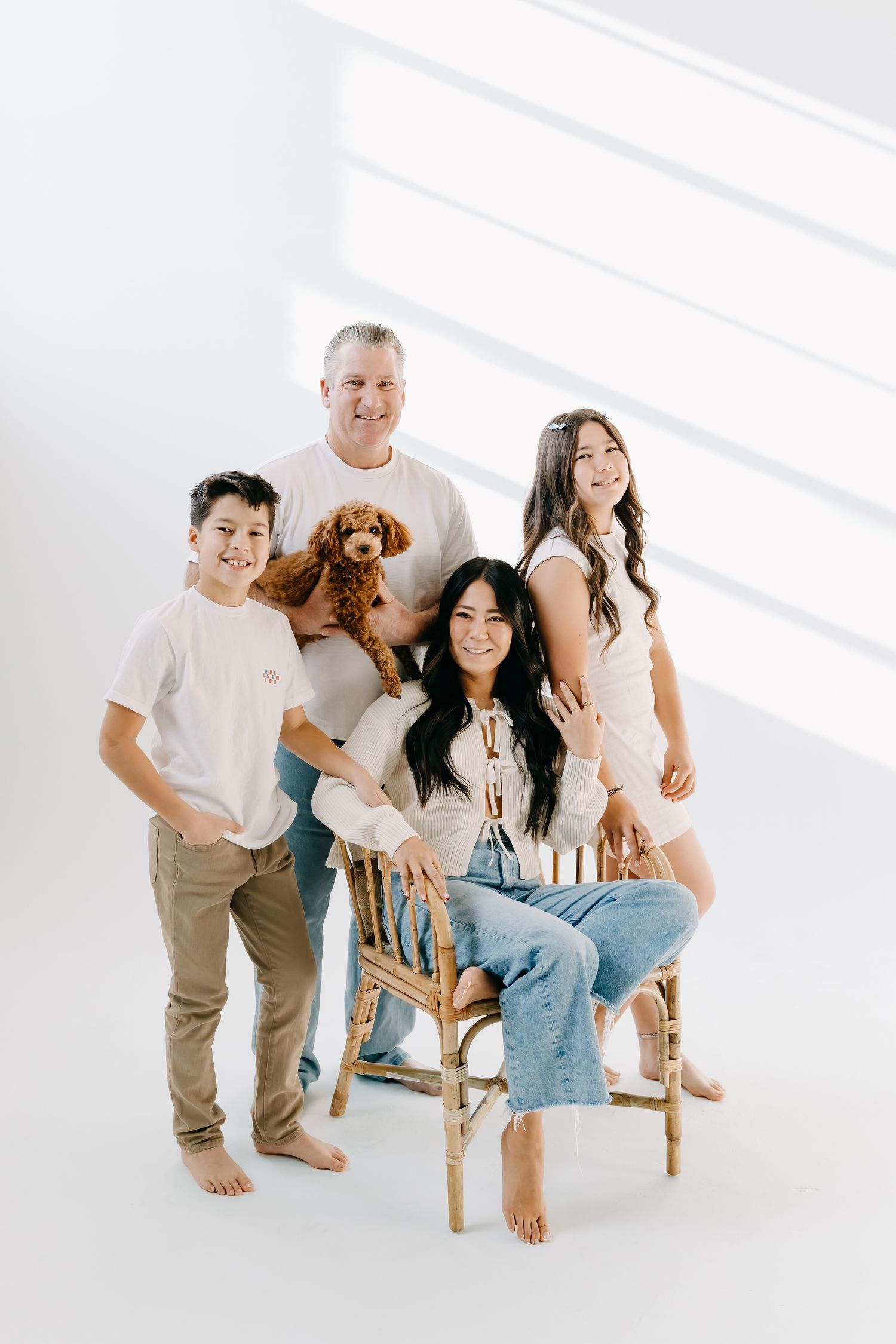 A family in white and denim poses playfully on a wooden chair with their brown dog in a bright minimalist setting.