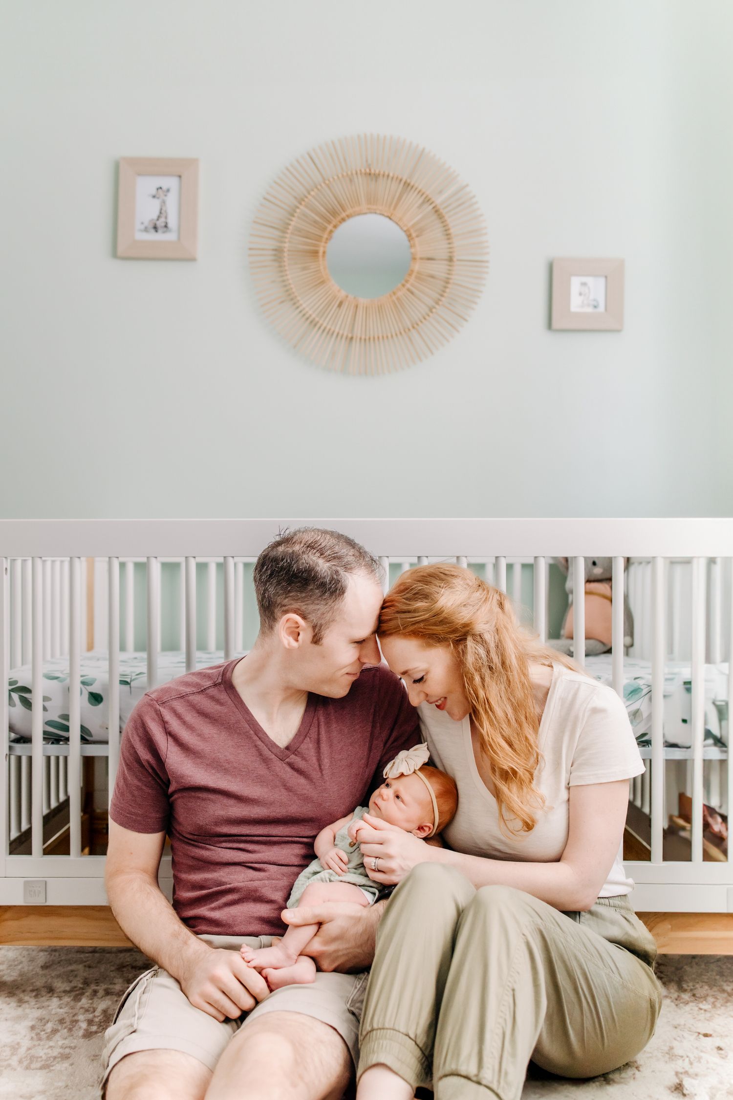 A couple sits on the floor near a white crib in a nursery decorated with sunburst mirrors and framed artwork.