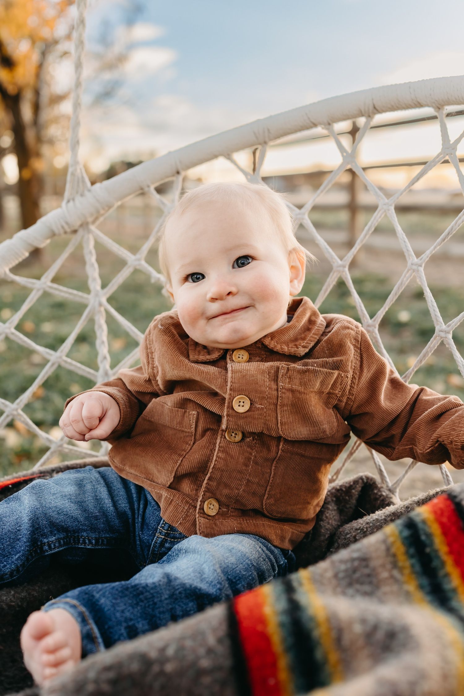 Baby wearing brown corduroy jacket and jeans sits on striped blanket outdoors on autumn day.