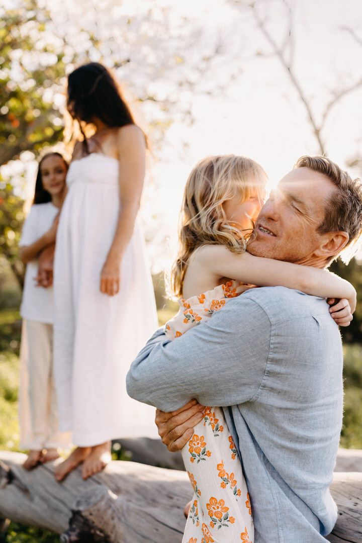 candid hug with father daughter captured in warm sunlight with people wearing white and floral dresses.
