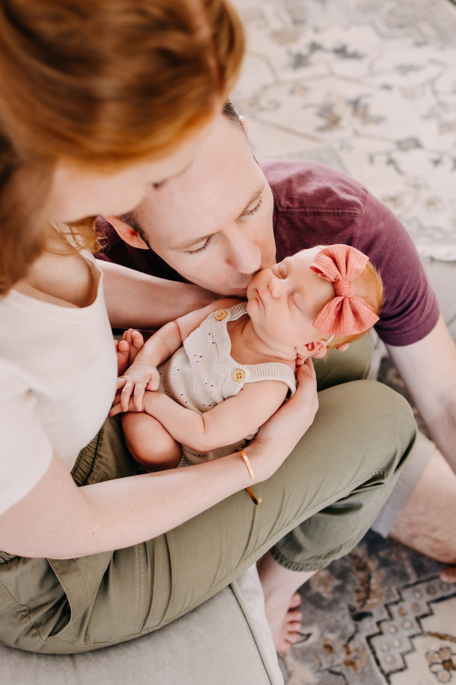 A tender family moment as parents share a loving interaction with their baby wearing a pink bow on a patterned rug.