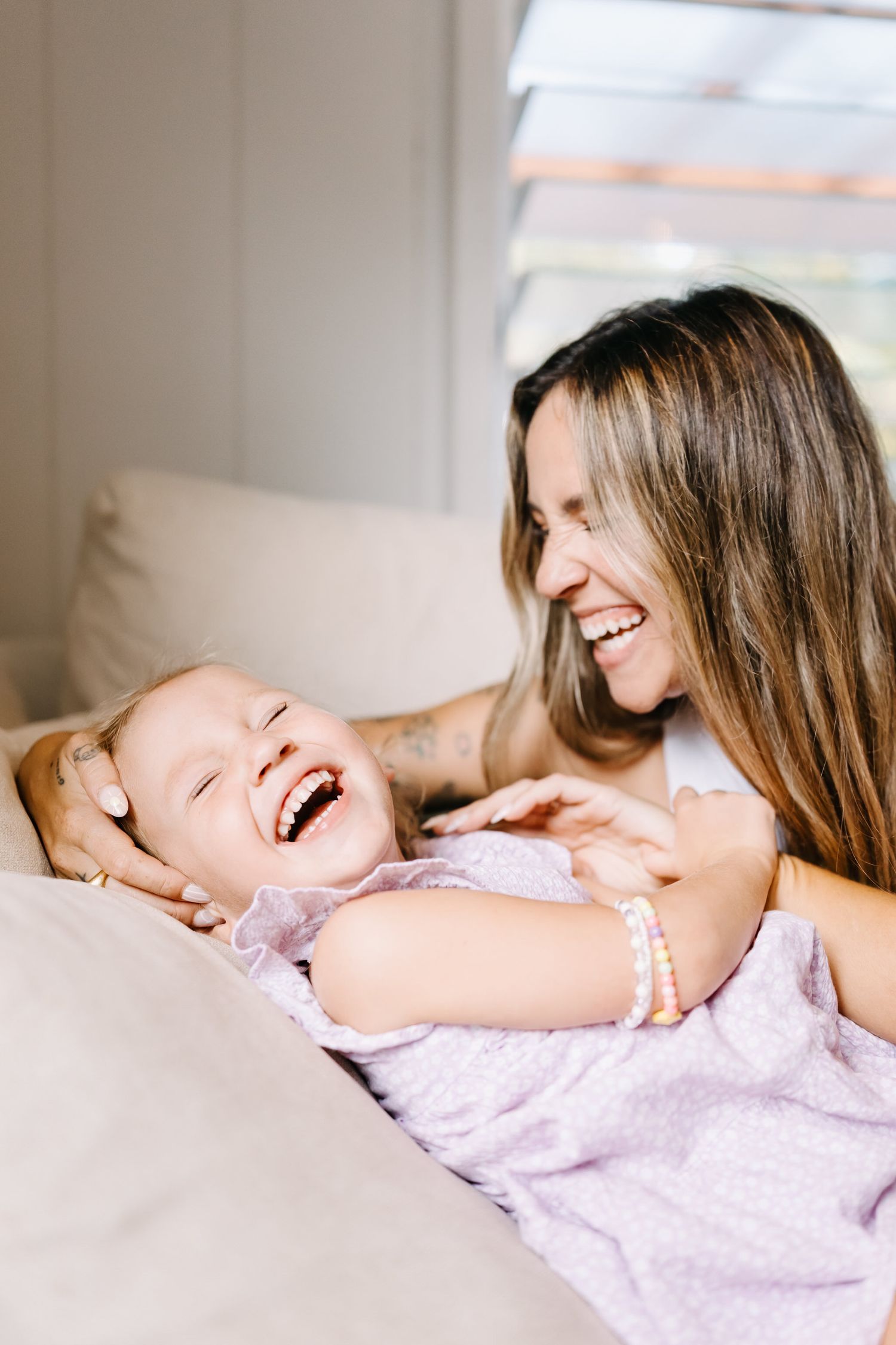 A joyful mother and daughter share laughter while lying together on a beige couch in natural lighting.
