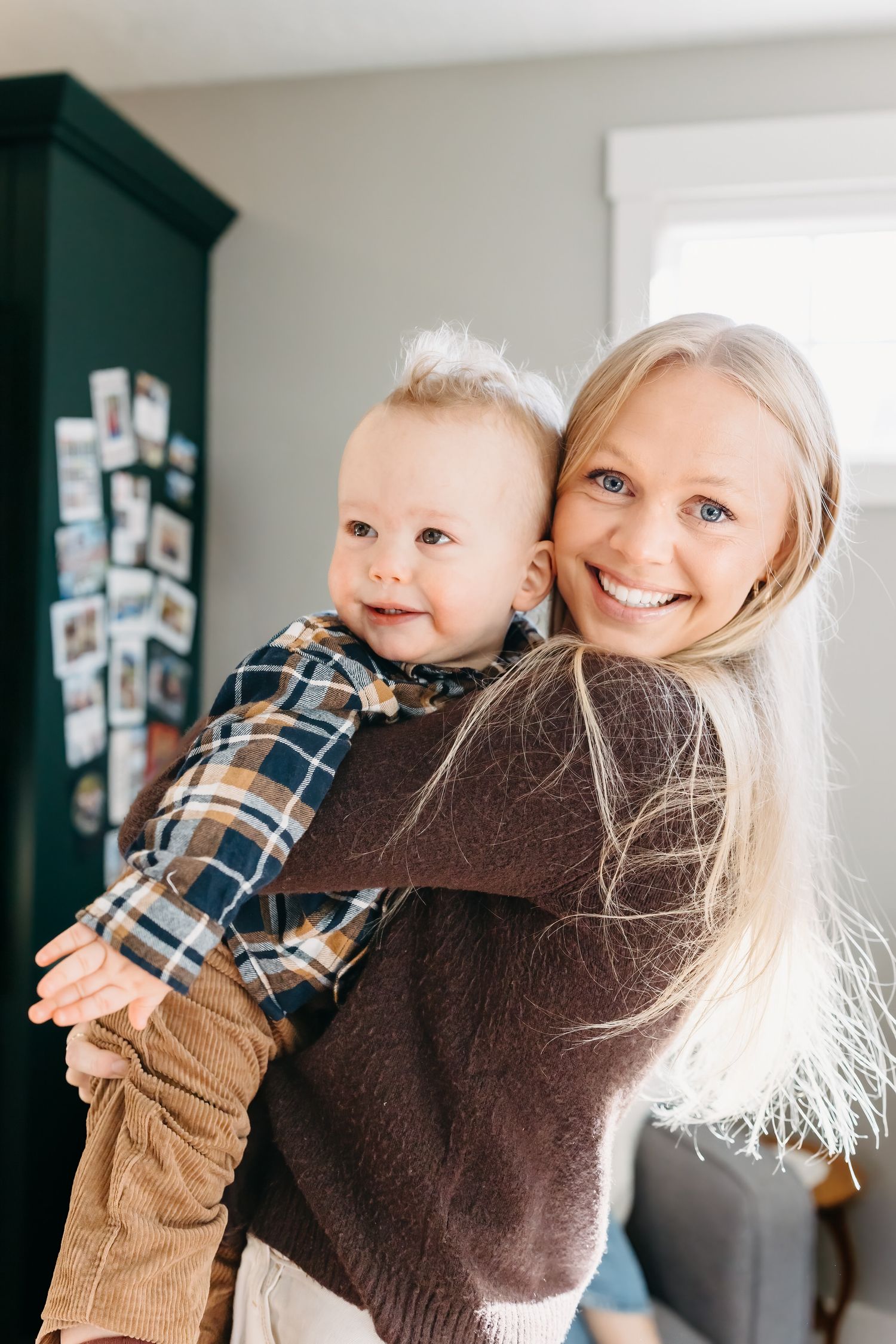 Aunt in a brown sweater holds a smiling nephew wearing a plaid shirt and khaki pants in a bright interior room.
