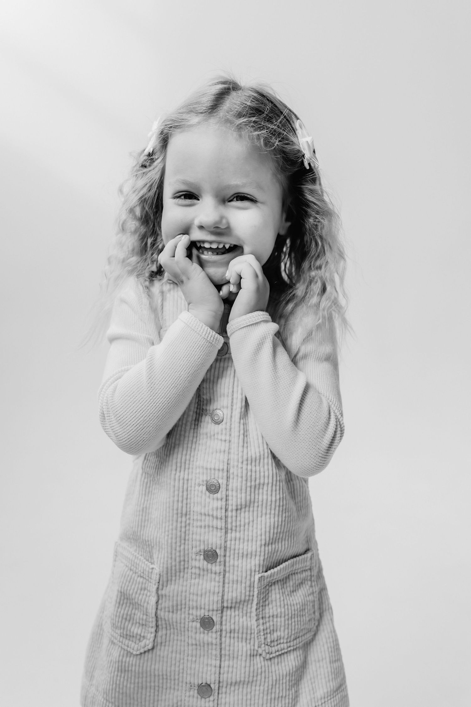 Black and white portrait of a child in a buttoned dress laughing with hands near face.
