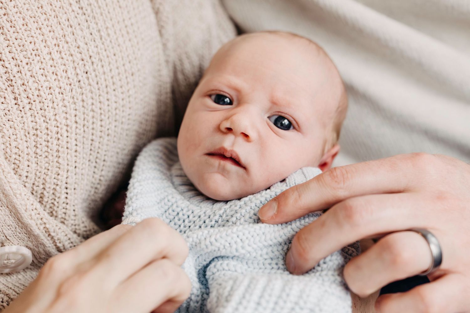 A baby wearing a light gray sweater lies on white bedding while being gently touched by an adult's hand.