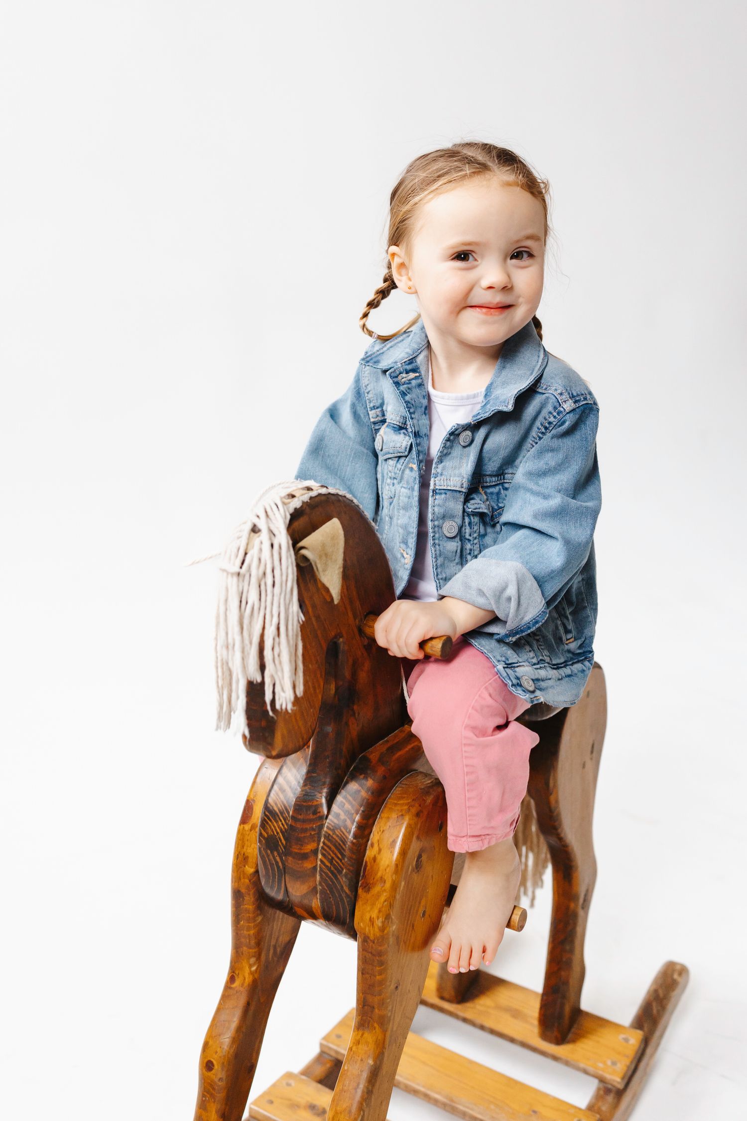 Child wearing denim jacket and pink pants sits on vintage wooden rocking horse toy against white background.