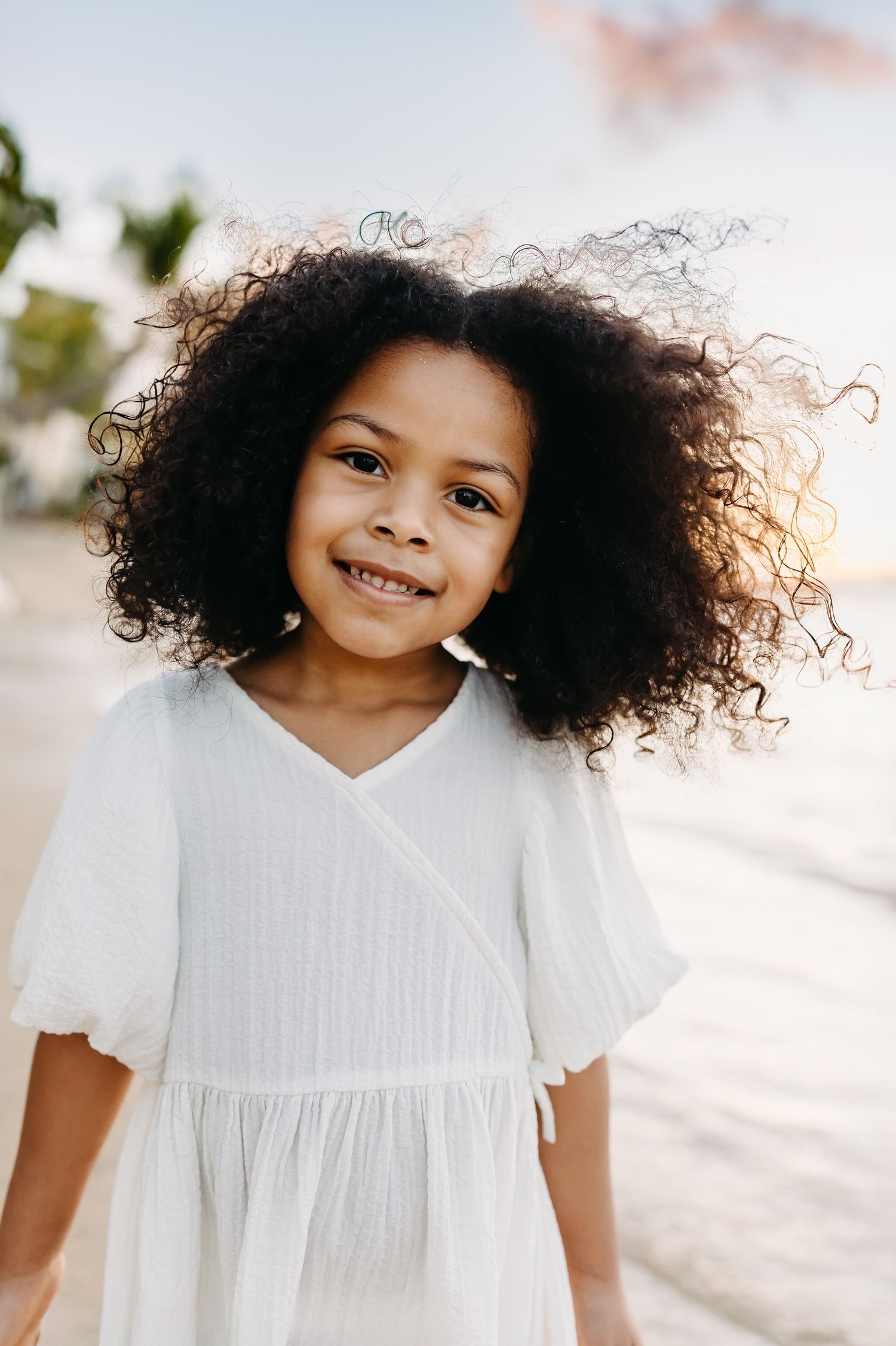 Young child wearing a white dress smiles while their curly hair flows in the breeze outdoors on a sunny day.