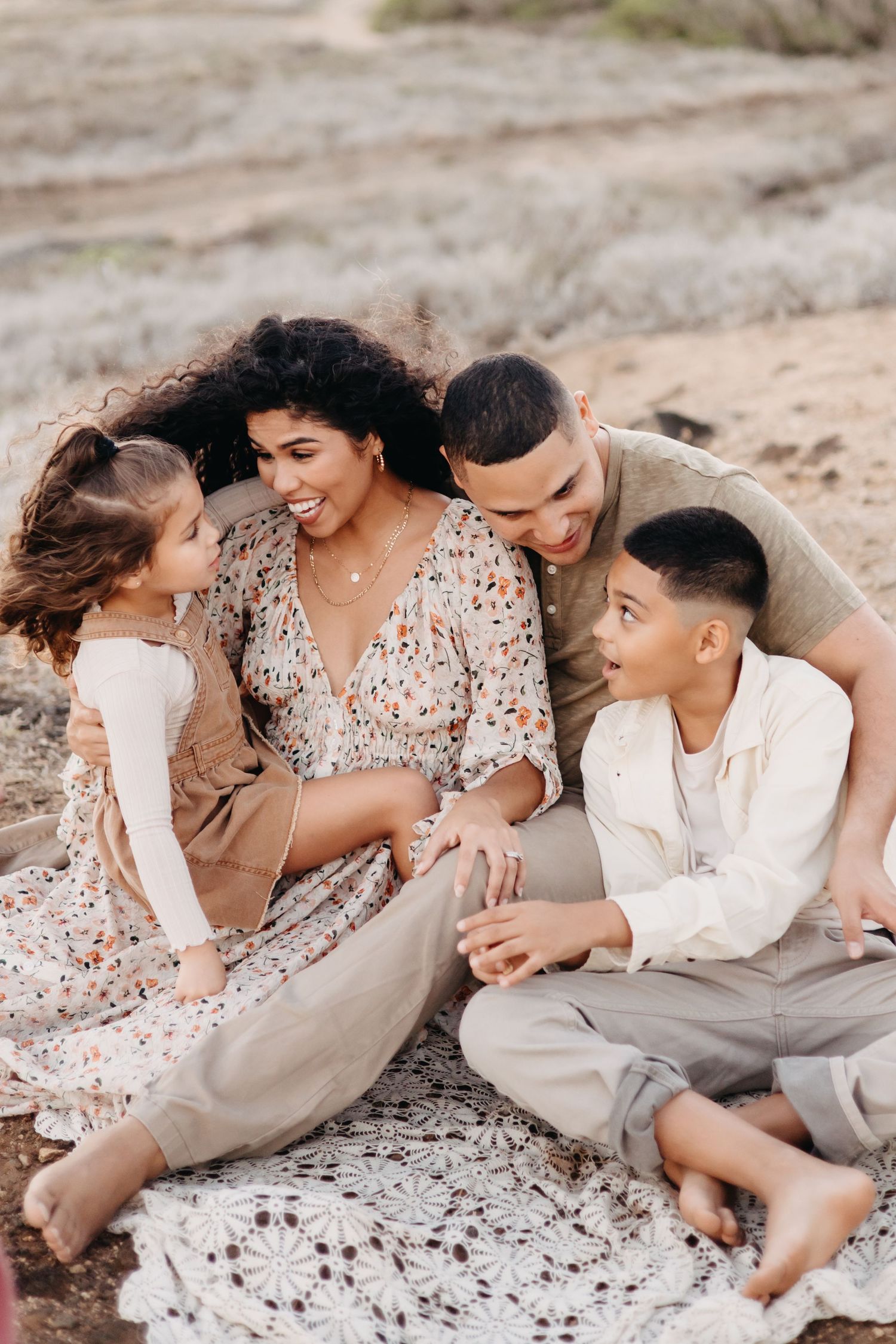 A romantic couple lounges together on a rocky beach in neutral toned outfits, sharing intimate moments in a photo series.