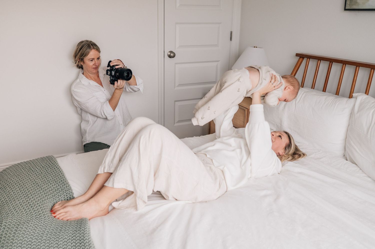 A person in white loungewear relaxes on a bed with white bedding in a light-filled bedroom.