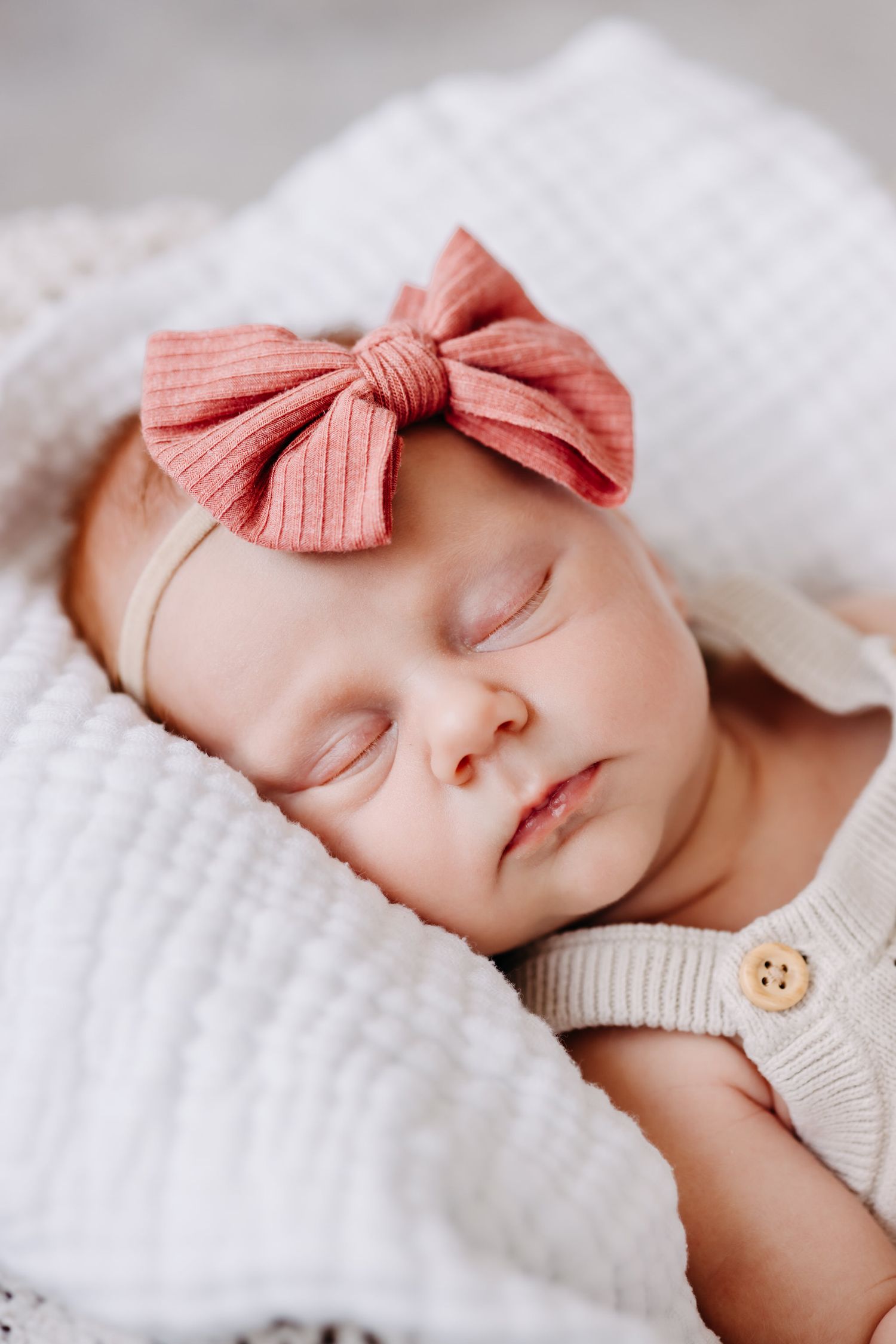 Sleeping newborn baby wearing white knit sweater and coral pink bow headband rests peacefully on white blanket.