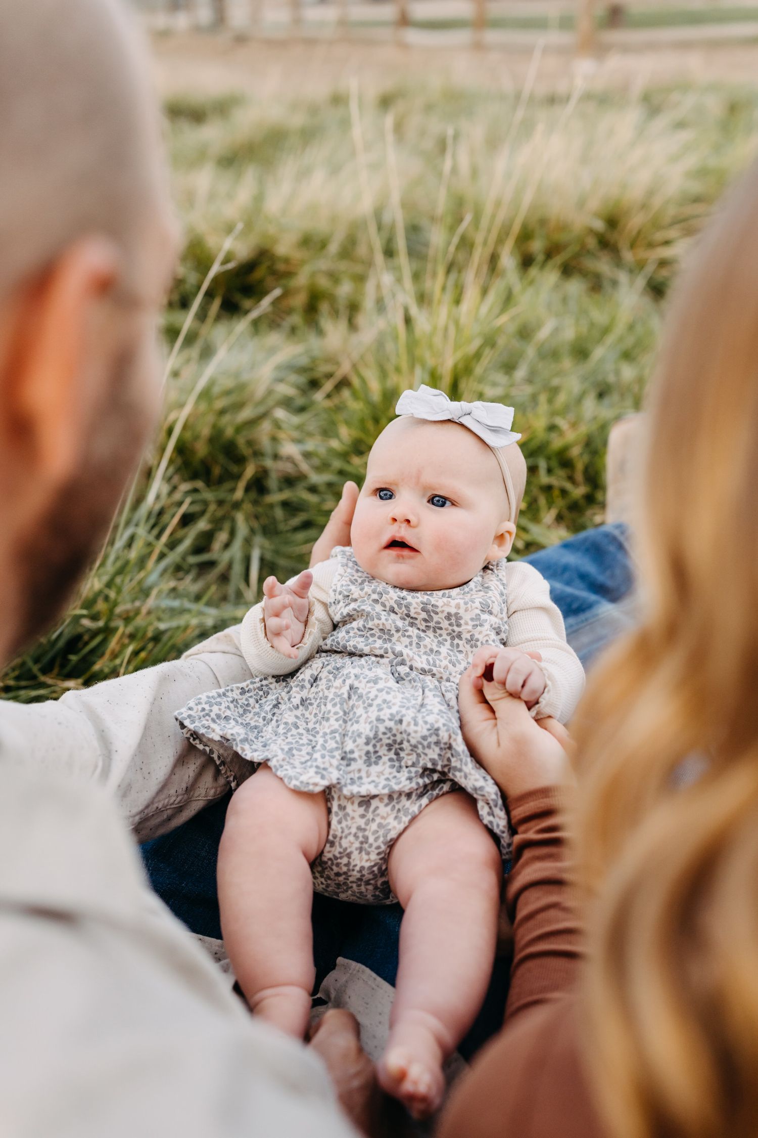Baby wearing a floral dress lies on grass while being held by parents in a series of candid outdoor shots.