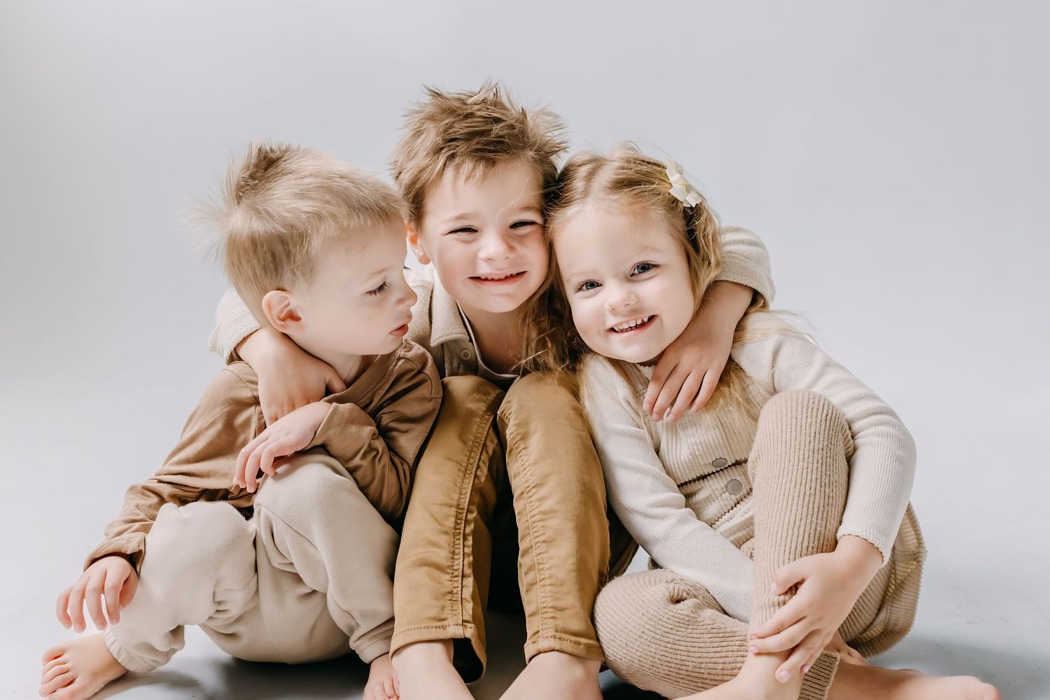 siblings in joyful embrace in matching beige and brown outfits against a white backdrop.