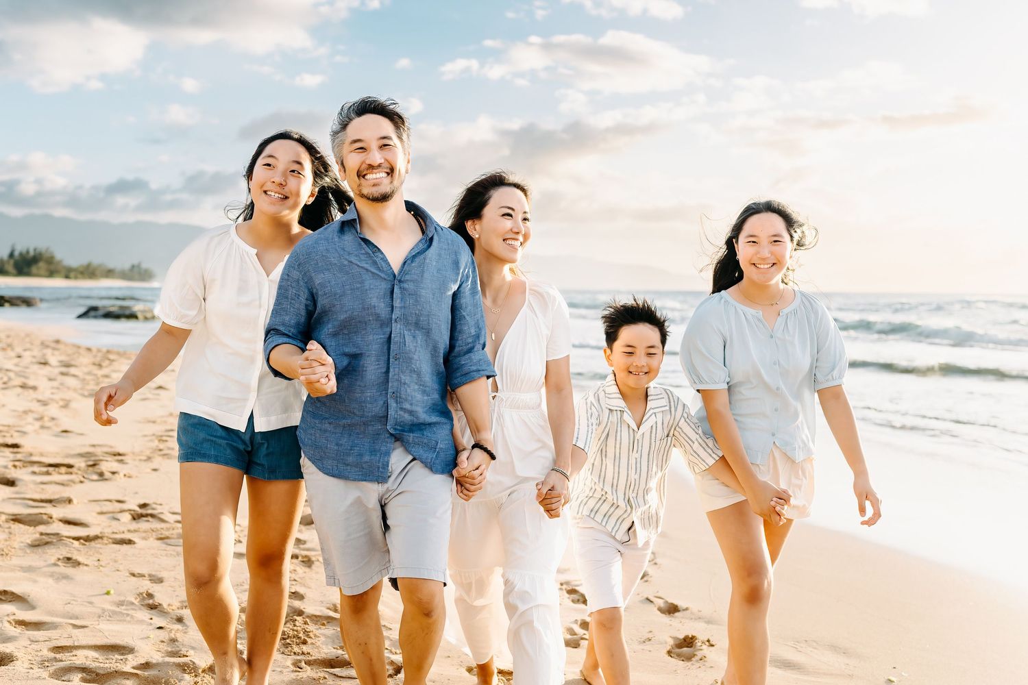 Family wearing coordinated blue and white outfits walking and laughing together on a sunny beach at sunset.