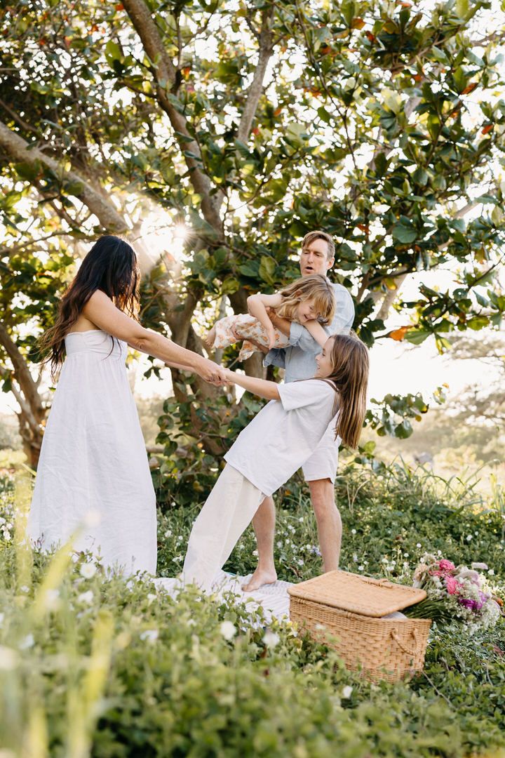 Family in white outfits having a picnic under a tree with a wicker basket and pink flowers in the background.