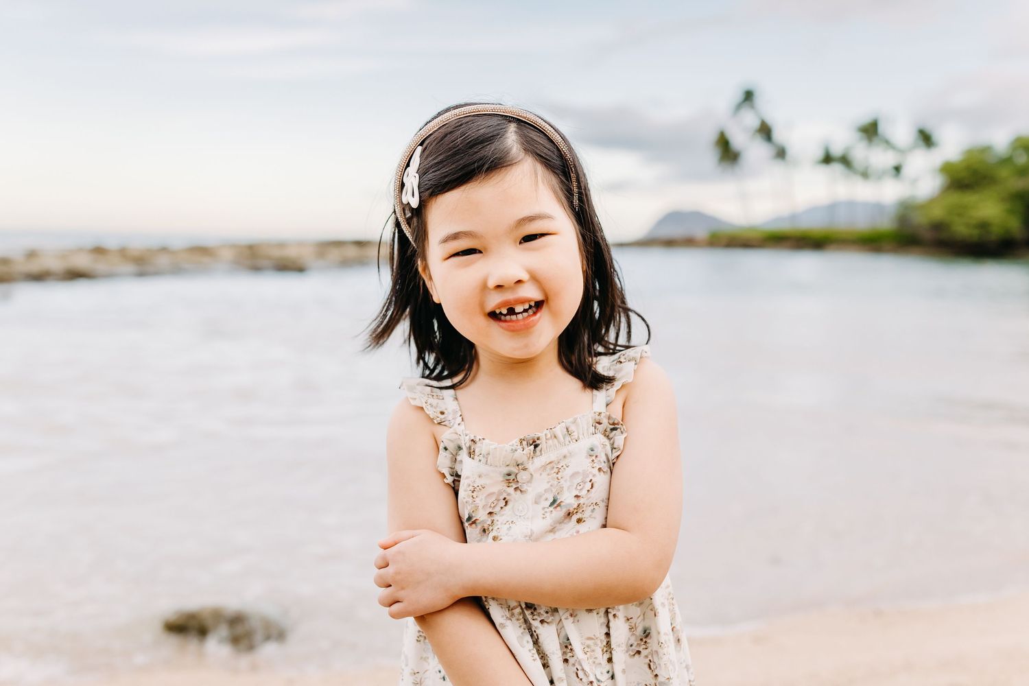 A child in a floral dress plays and laughs joyfully on a sunny beach with ocean waves in the background.