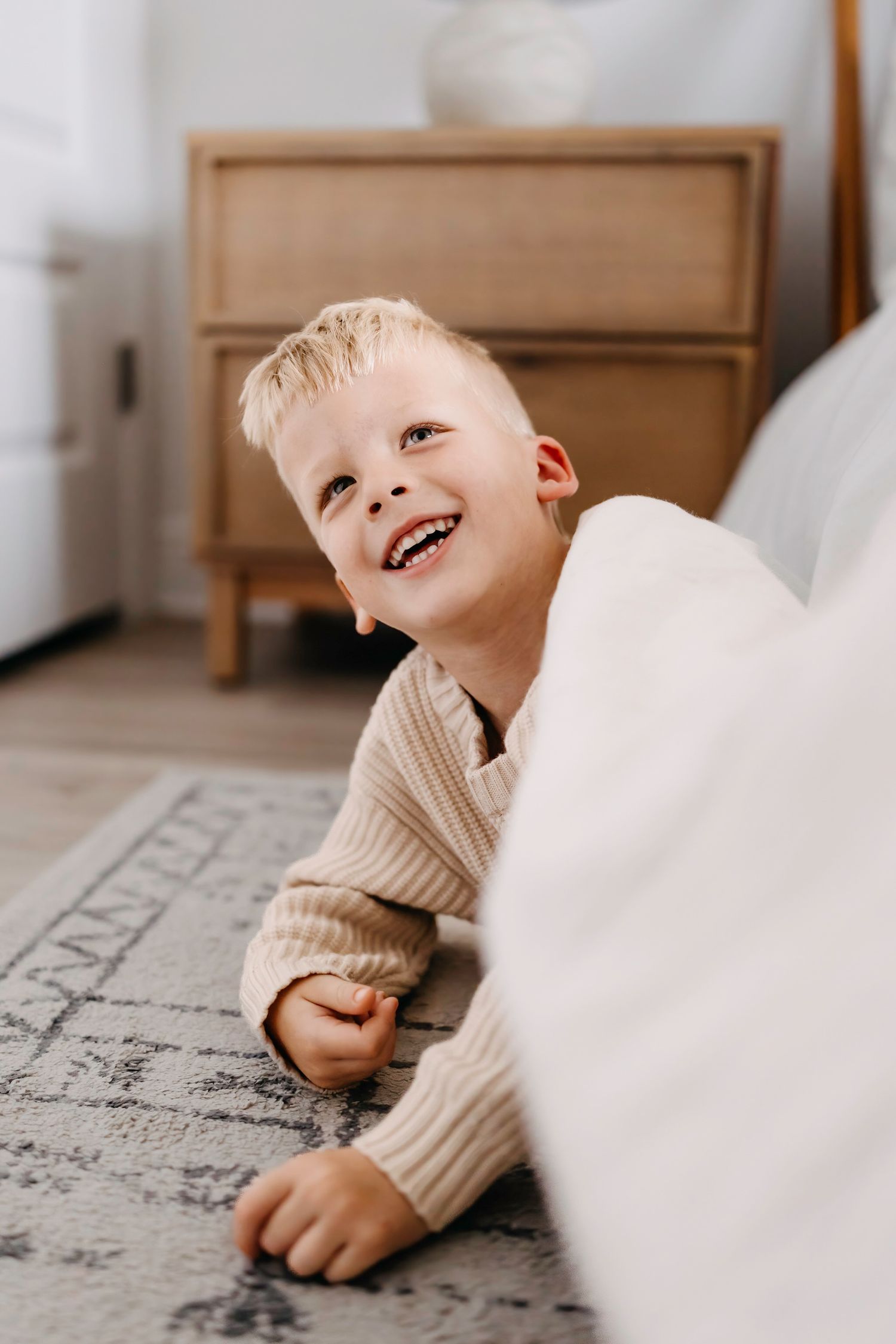 A young child in a cream sweater lies on a patterned rug, laughing joyfully with a bright smile.