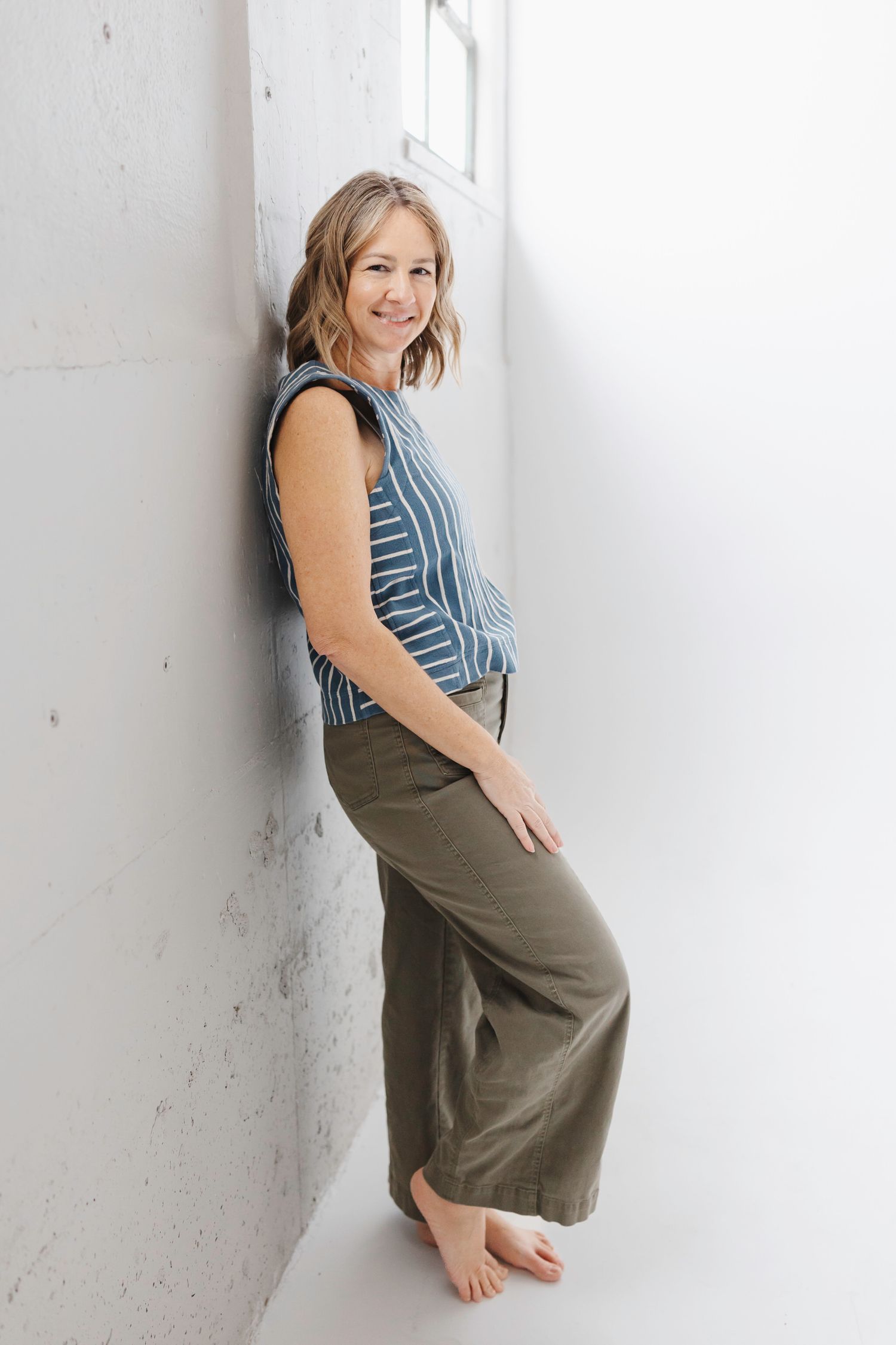 Woman in striped sleeveless top and olive pants poses barefoot against white wall in natural lighting.