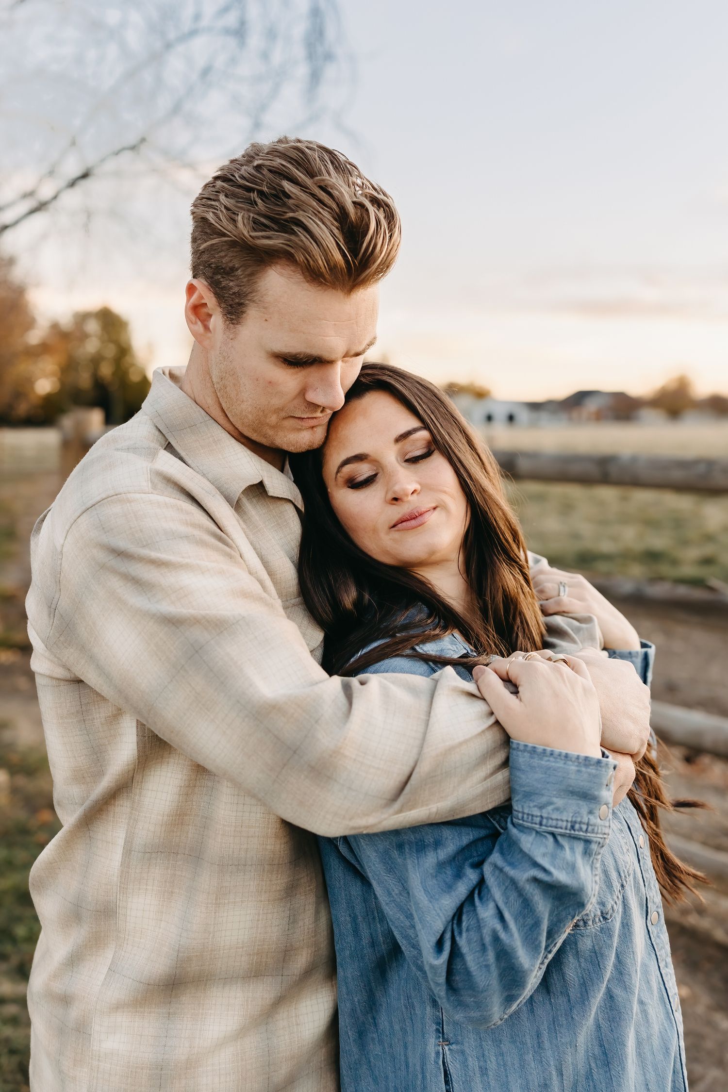 A couple embraces tenderly in warm sunlight while wearing casual denim and beige clothing during an outdoor photoshoot.