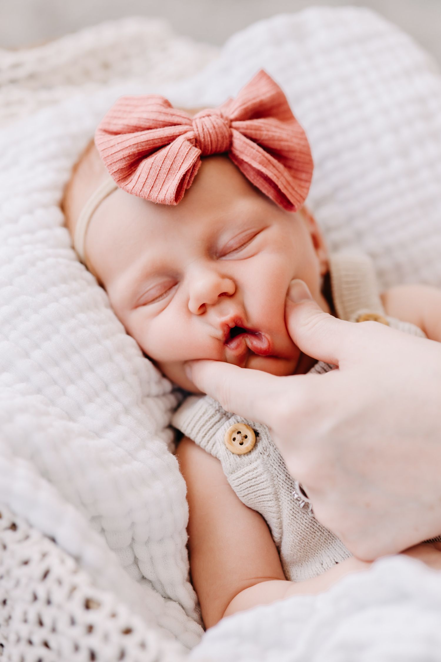 A sleeping newborn baby wearing a pink bow headband rests peacefully on white textured bedding.
