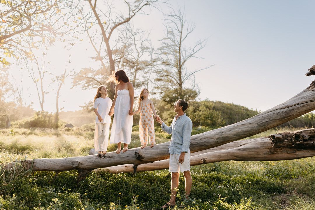 A family is white, creams, and blue clothing poses on a fallen tree trunk during golden hour in a scenic outdoor setting.