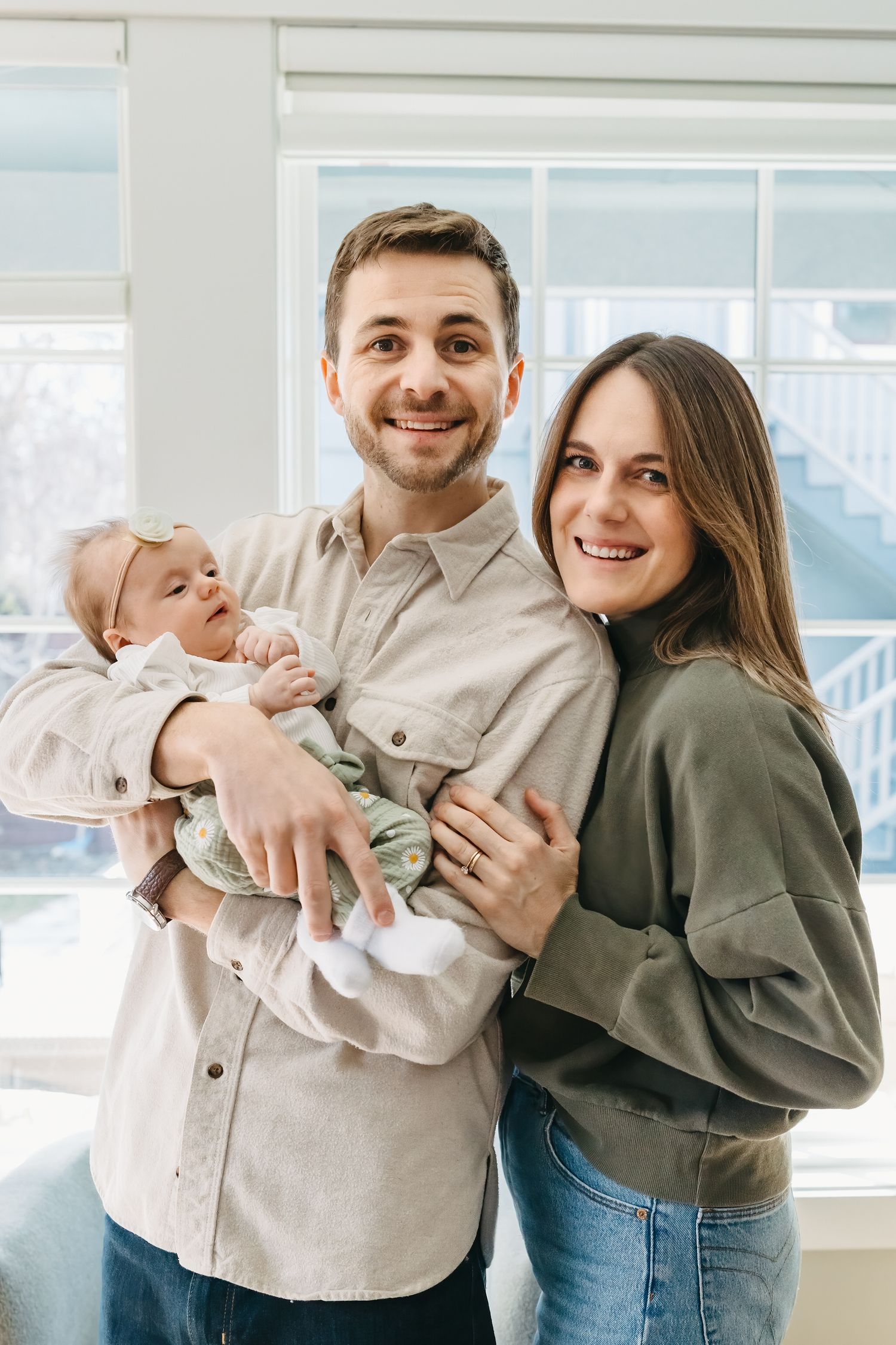 A couple in casual attire smiles while holding a baby in a bright, sunlit room with windows.