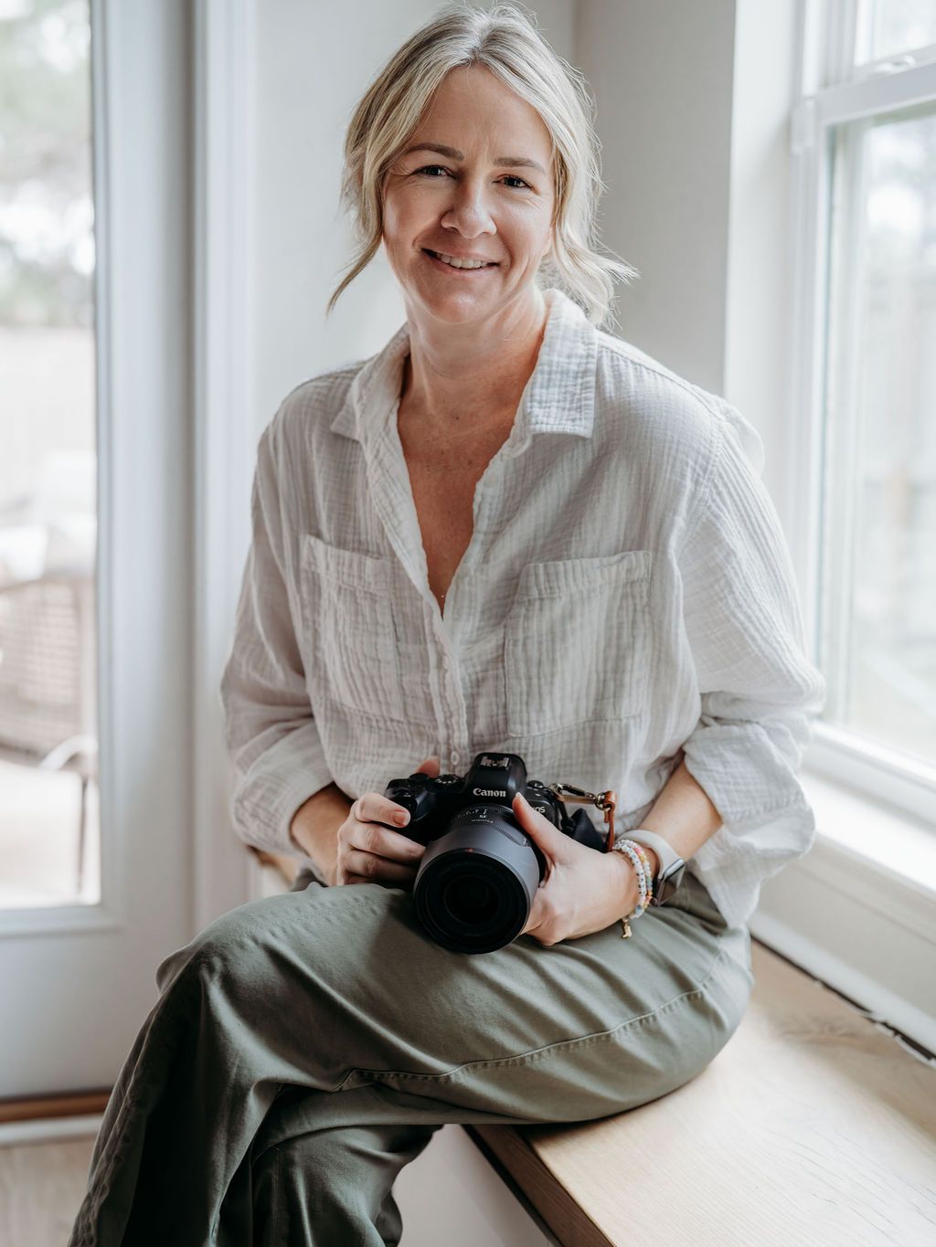 Professional photographer in white shirt sits by window while reviewing images on digital camera.