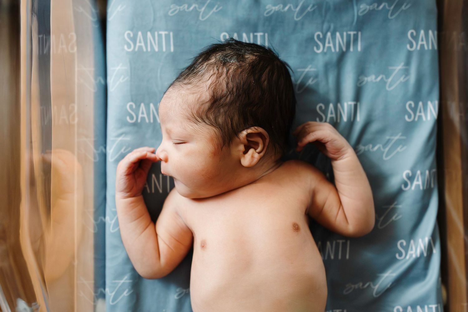 Sleeping newborn baby lying on blue patterned fabric with 'Santi' text repeated in background.