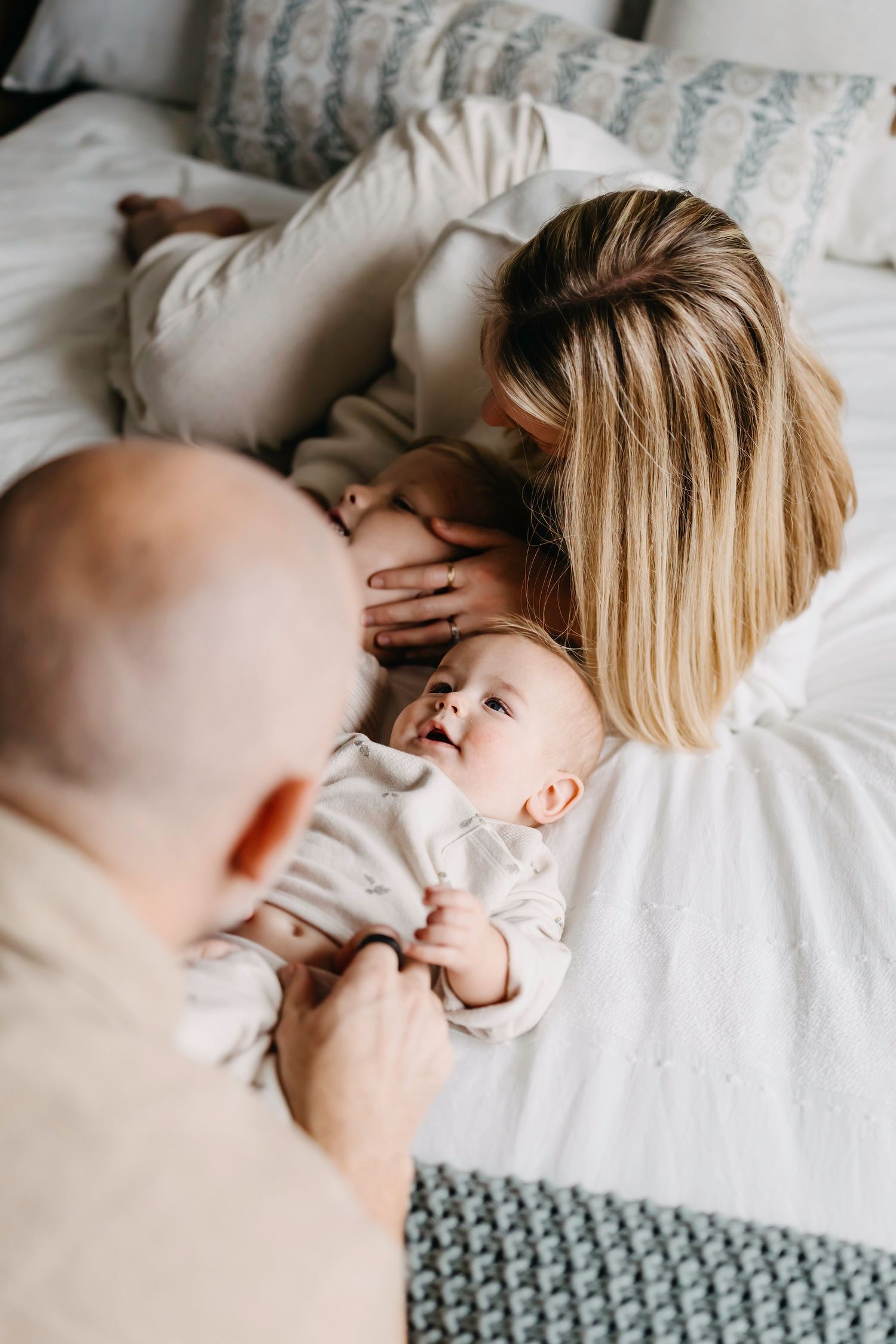 A tender family moment captured from above as they lay together on white bedding with a knitted blanket visible.