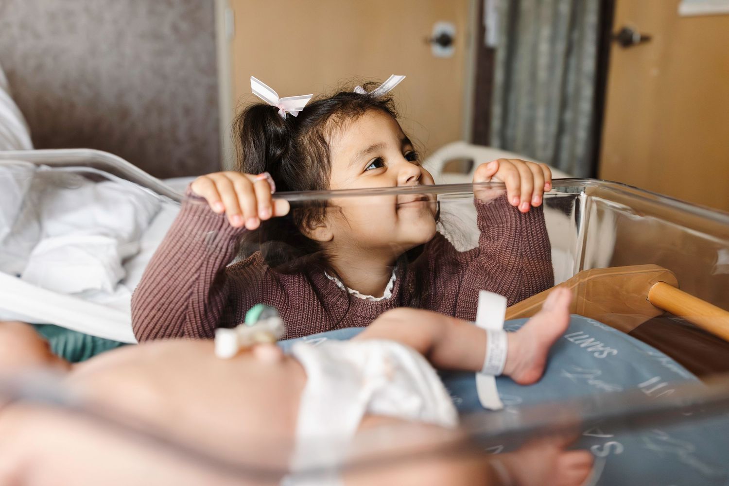 Child playfully makes faces while visiting new sibling in a hospital bassinet.
