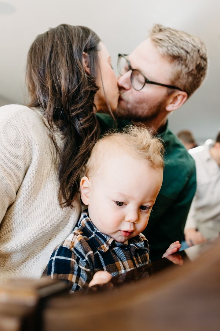 A small toddler in a plaid shirt plays on a piano while parents kiss in the background