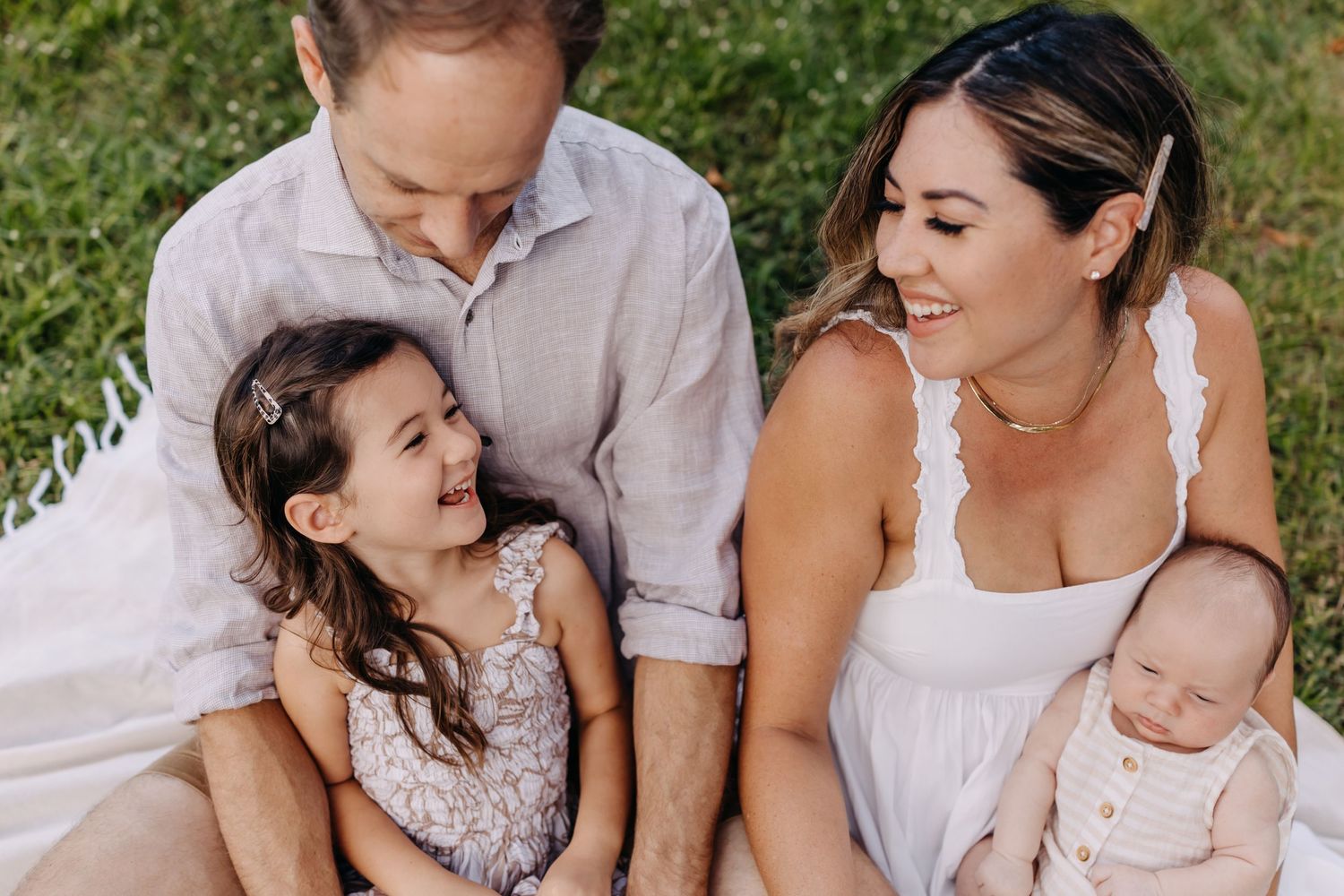 A family sits together outdoors wearing white and cream colored clothing while sharing a joyful moment.