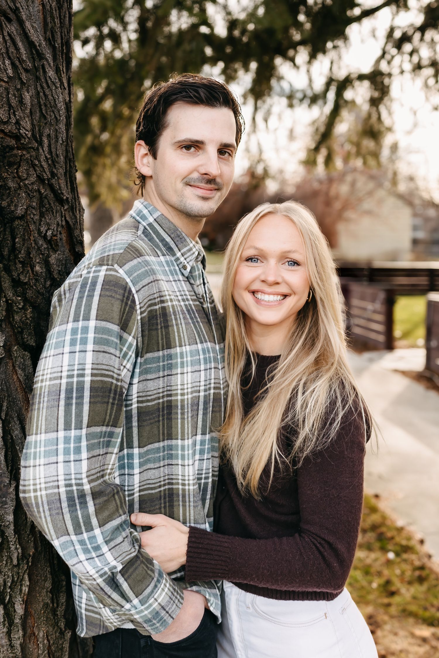 A couple shares romantic moments outdoors in autumn, wearing plaid and brown sweaters while embracing near trees.