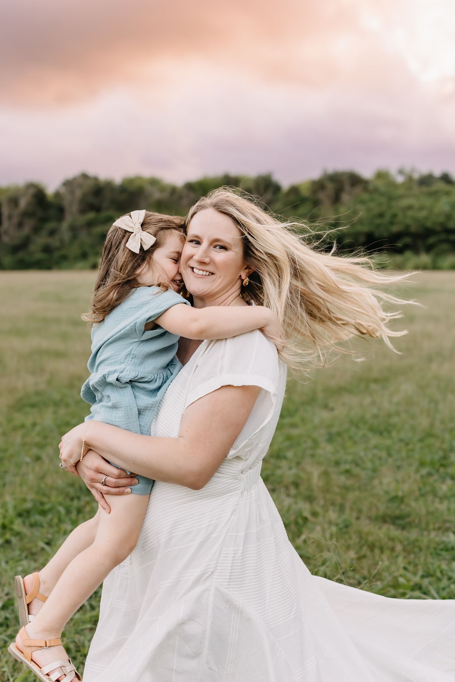 A joyful moment captured outdoors as a mother in a white dress spins and holds her young child in a grassy field.