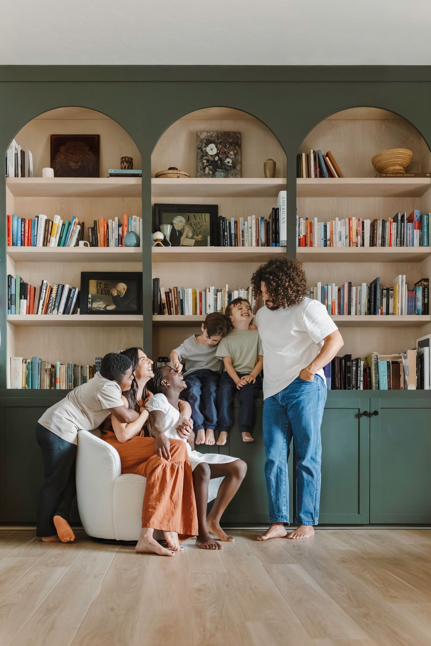 Family relaxing together on floor in front of built-in bookshelves with green cabinets in modern living room.