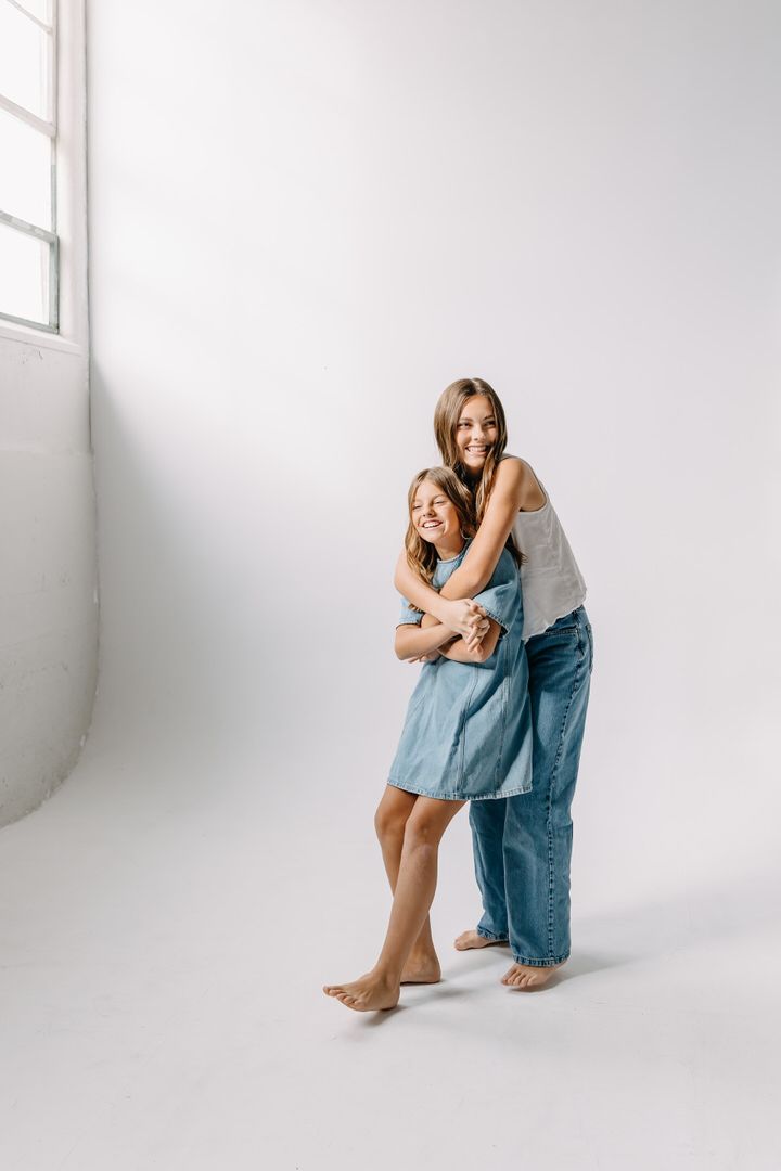Two people in casual denim and white outfits embrace while standing against a minimalist white studio background.