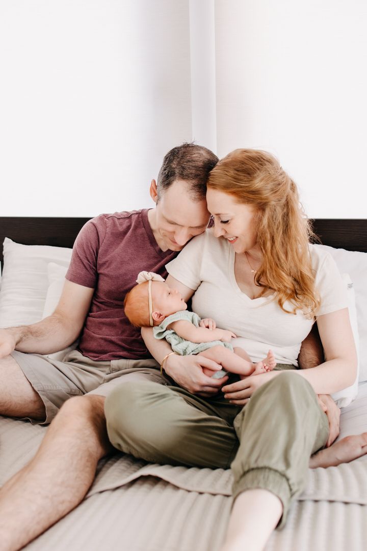 A couple relaxes on a white bed while lovingly cradling and bonding with their newborn baby.