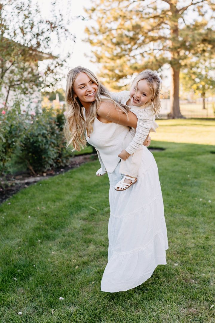 Mother and daughter in white dresses playing and twirling together on green grass during golden hour sunset.