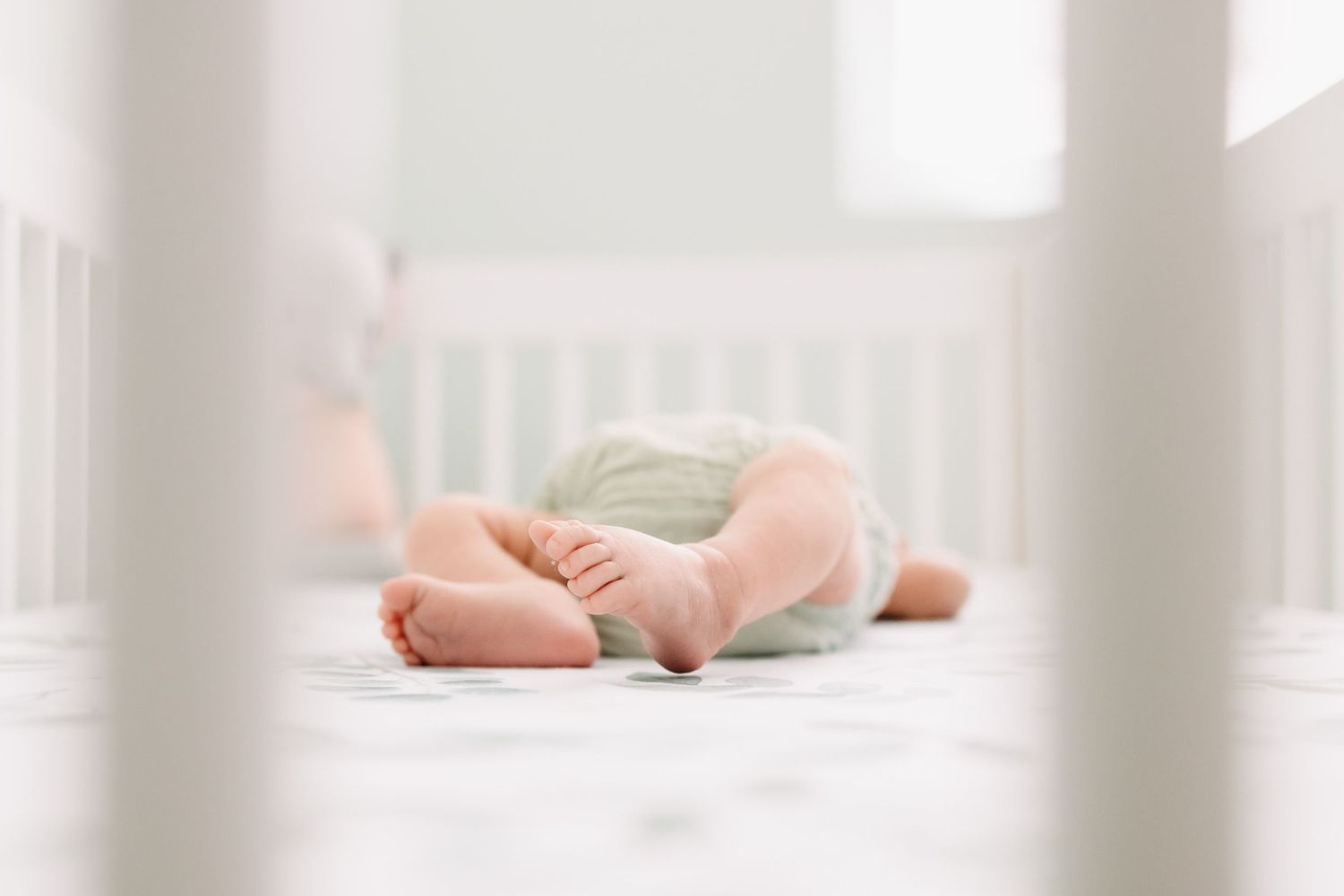 Tiny baby feet peek through white crib bars in a soft, dreamy nursery setting.
